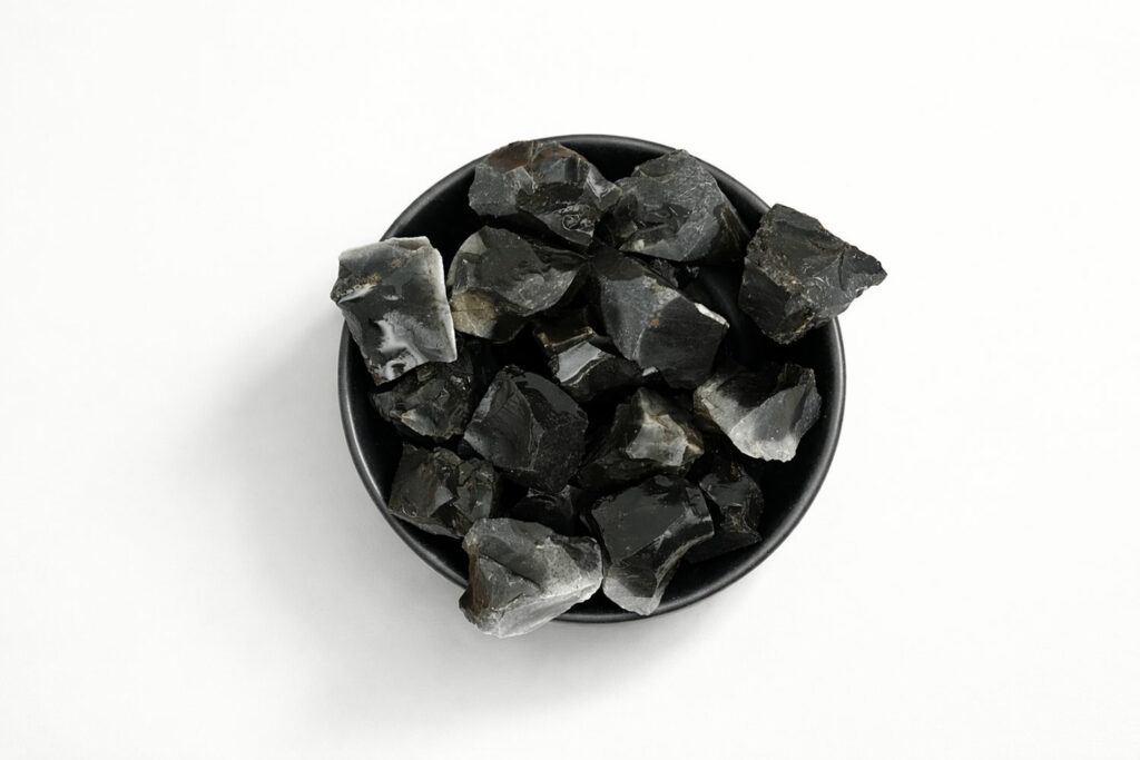 Bol noir rempli de morceaux de pierre onyx noire brute naturelle de différentes tailles sur un fond blanc.